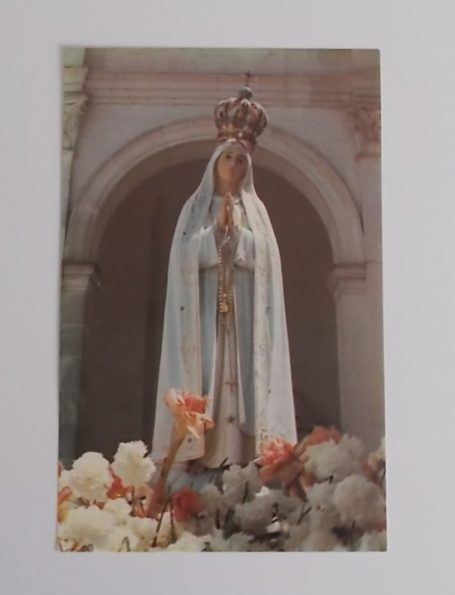 Statue der Jungfrau Maria, umgeben von weißen Rosen, stehend vor einem Bogen.