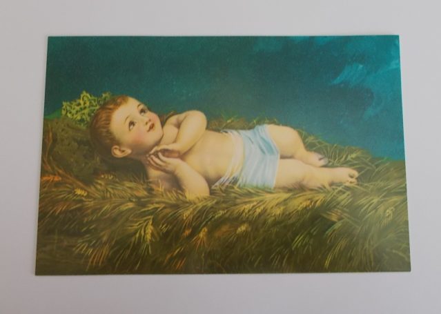 Postkarte "Jesuskind von Betlehem" Ein lächelndes Kind liegt auf grünem Gras mit einem blauen Himmel im Hintergrund.