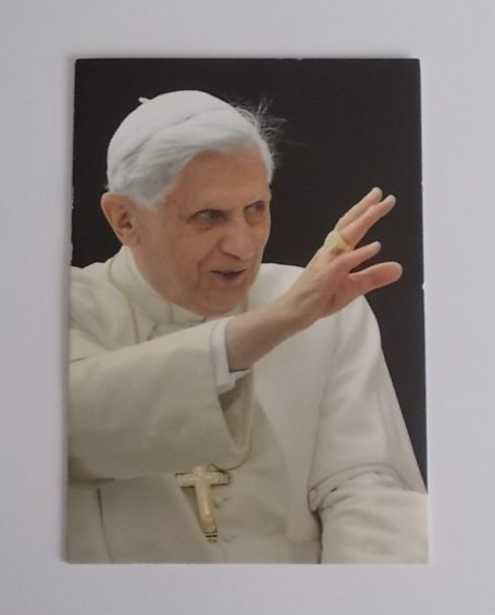 Papst Benedikt XVI. in weißer Kleidung, mit erhobener Hand.